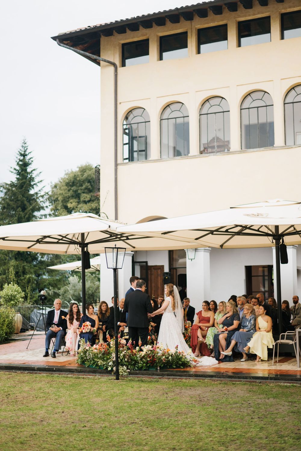 Wedding guests gather under white umbrellas on a lawn outside a Mediterranean-style venue.