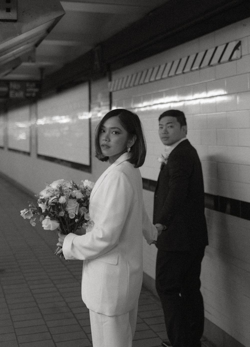 A wedding couple stands in a white-tiled subway station corridor creating artistic black and white photographs.