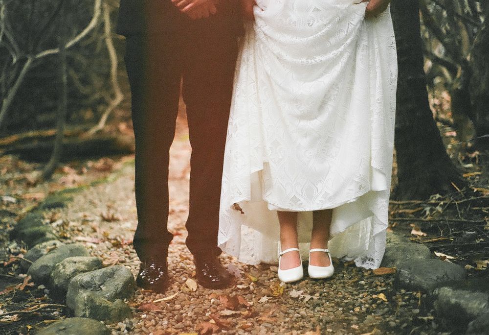 Closeup of feet and white dresses walking on a forest path during a rustic outdoor wedding celebration.