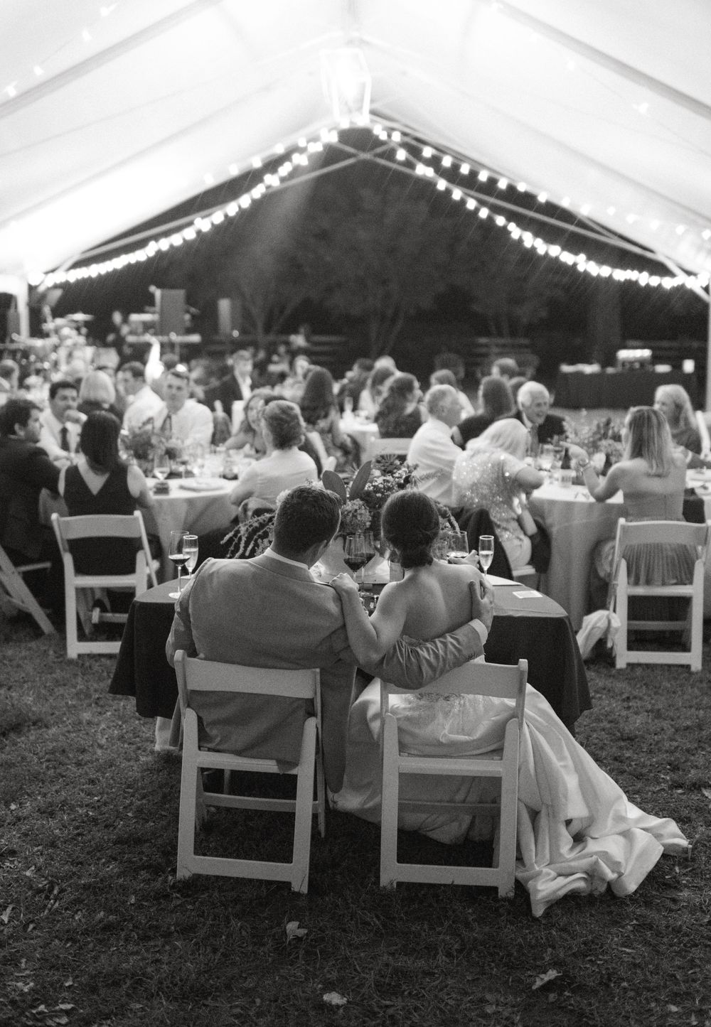 Wedding guests seated at tables under a tent with string lights during an outdoor reception celebration.