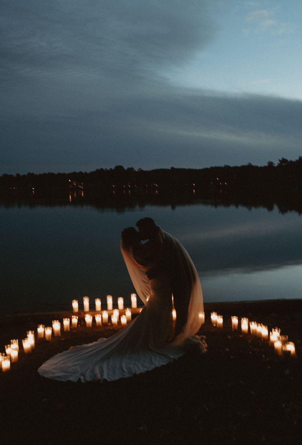 A romantic lakeside scene at dusk with glowing candles lining the shore and a silhouette in an elegant dress.