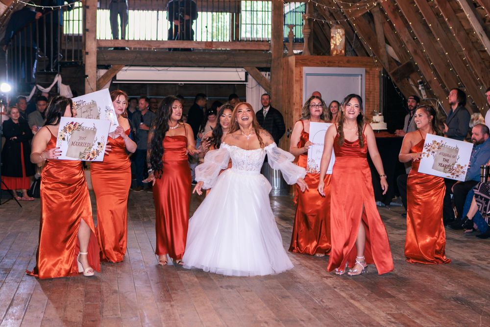 Bridal party in white wedding gown and orange bridesmaid dresses pose together in rustic wooden barn venue.