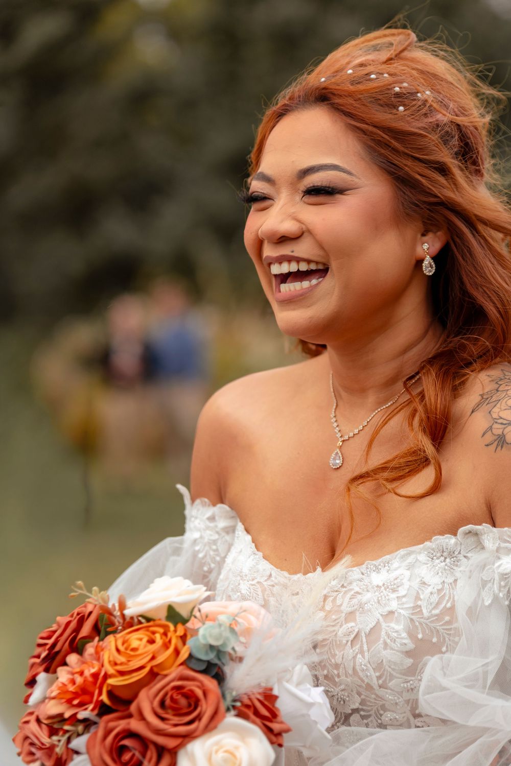 A joyful bride in a white lace wedding dress holds an autumn-colored bouquet of orange and red roses during an outdoor ceremony.
