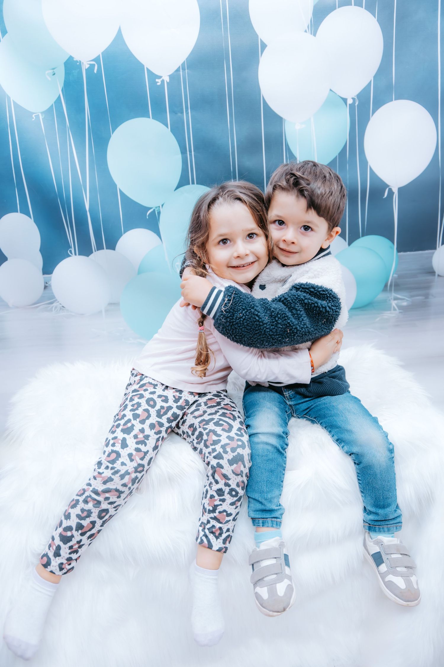 Two children hugging and smiling in front of white and blue balloons against a blue backdrop.