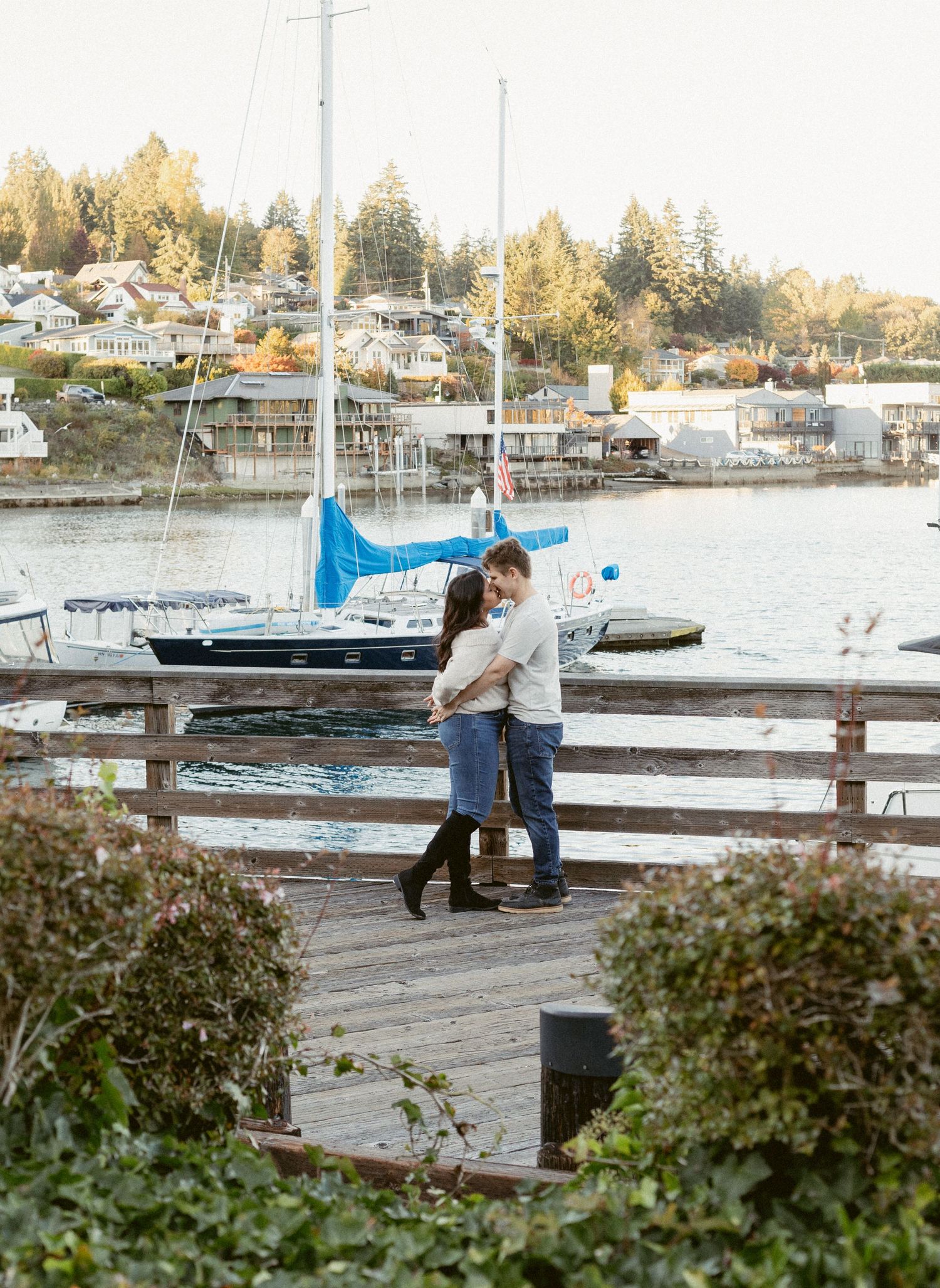 A couple walks along a wooden dock at sunset with boats and autumn trees in the marina background.