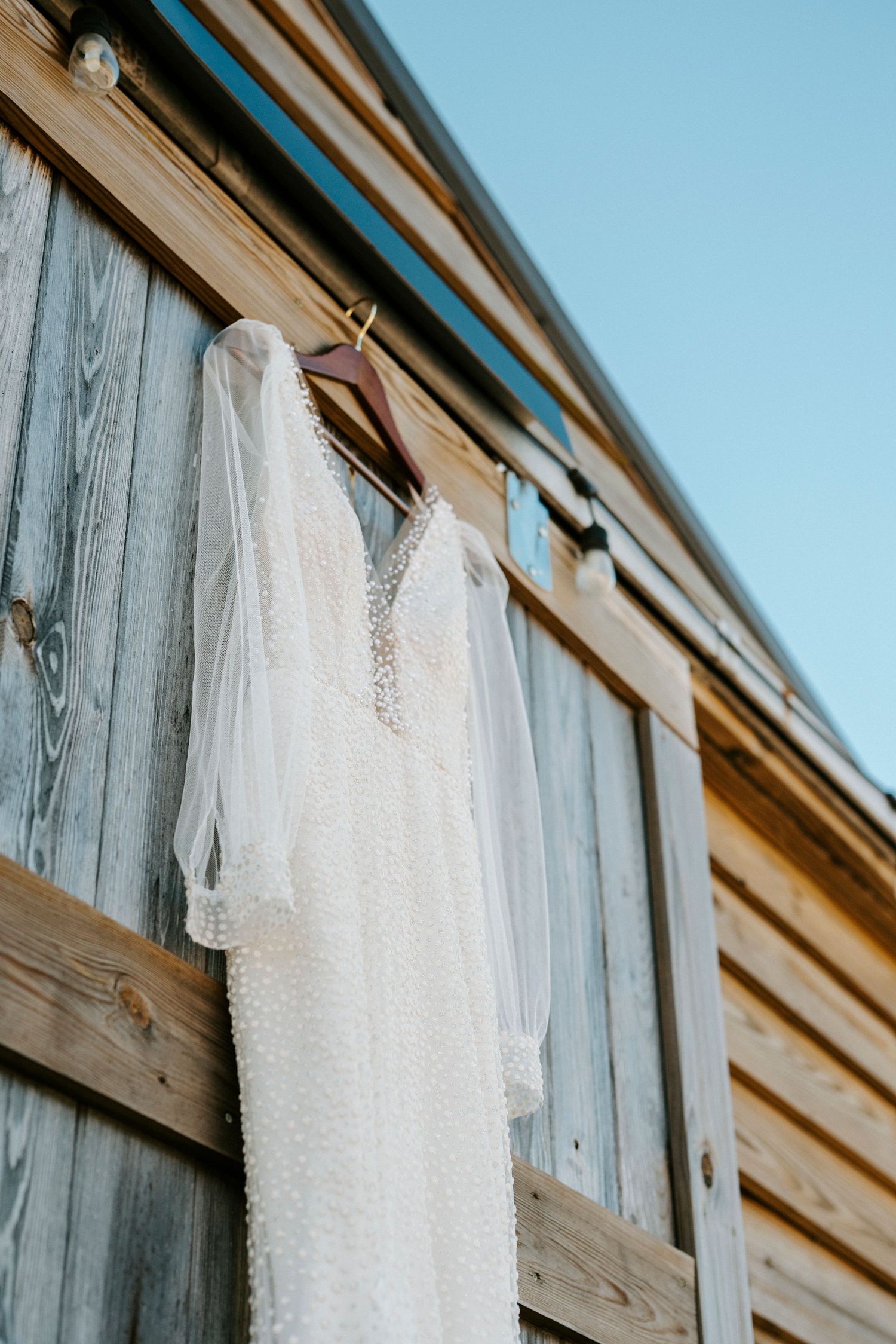 A delicate white lace wedding dress hangs on a rustic wooden barn wall against a blue sky.
