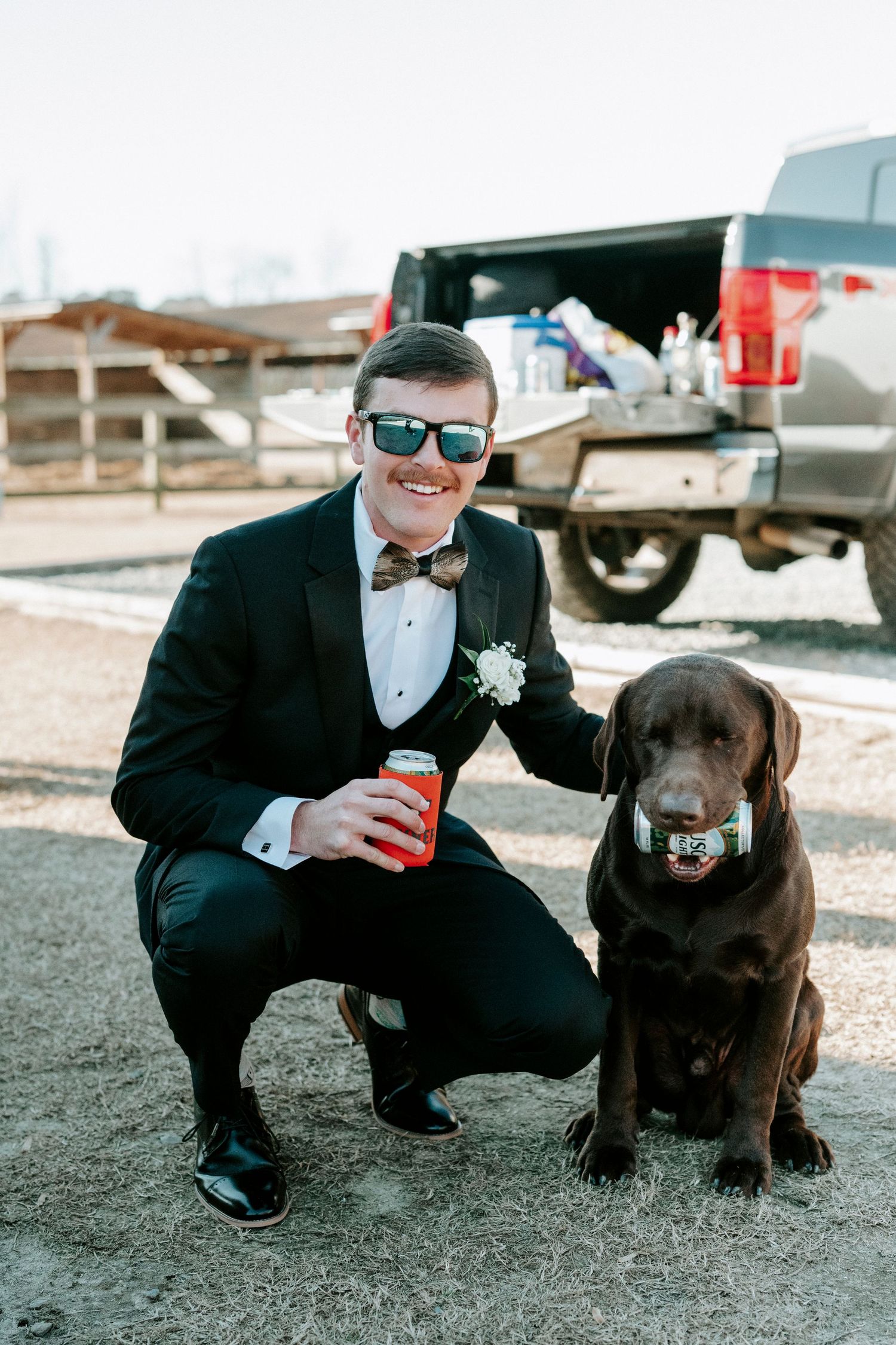 A groomsman in a tuxedo shares a playful moment with a chocolate Labrador retriever wearing a bow tie.