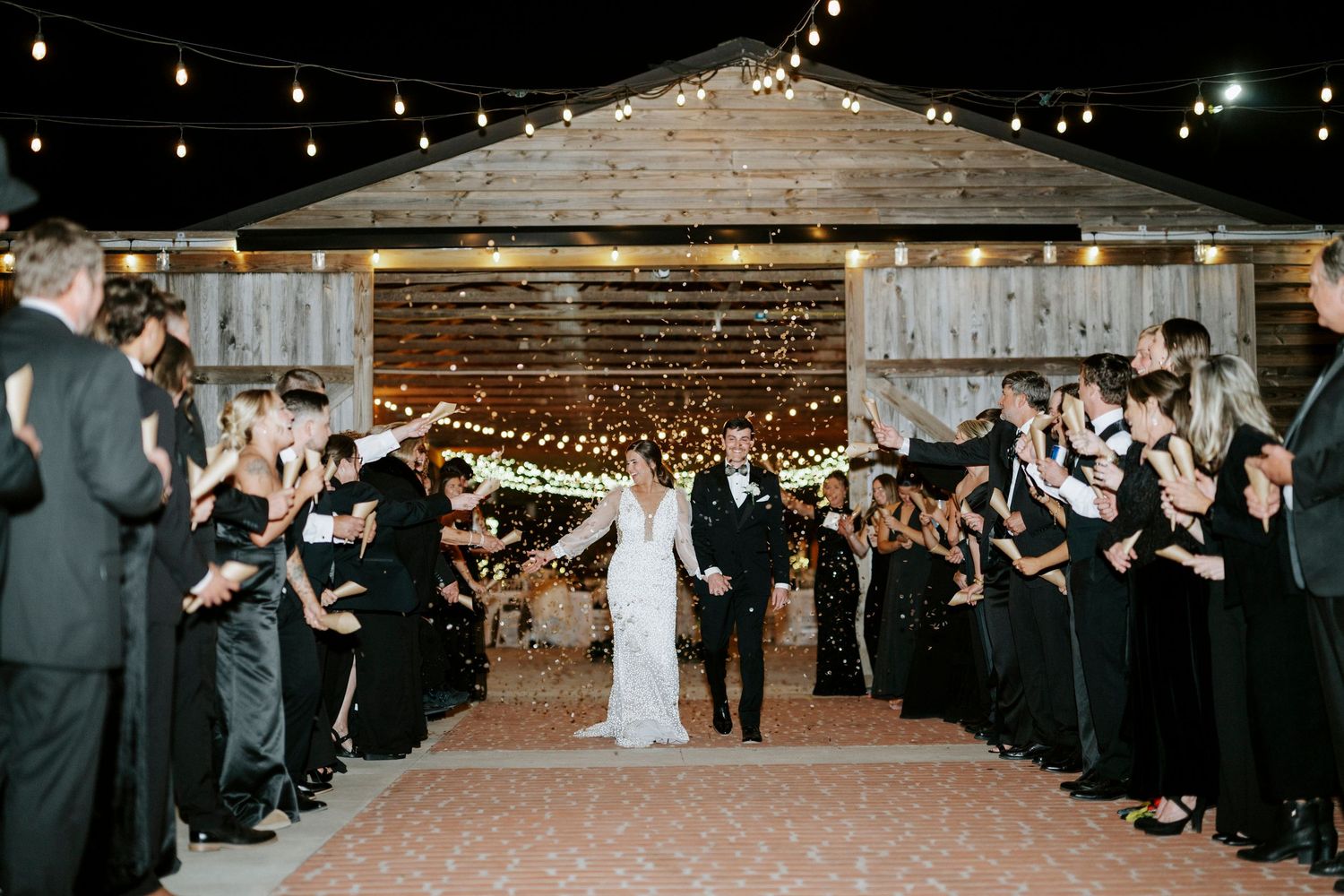 Wedding guests form a sparkler exit tunnel at night as couple walks through rustic barn venue with string lights overhead.