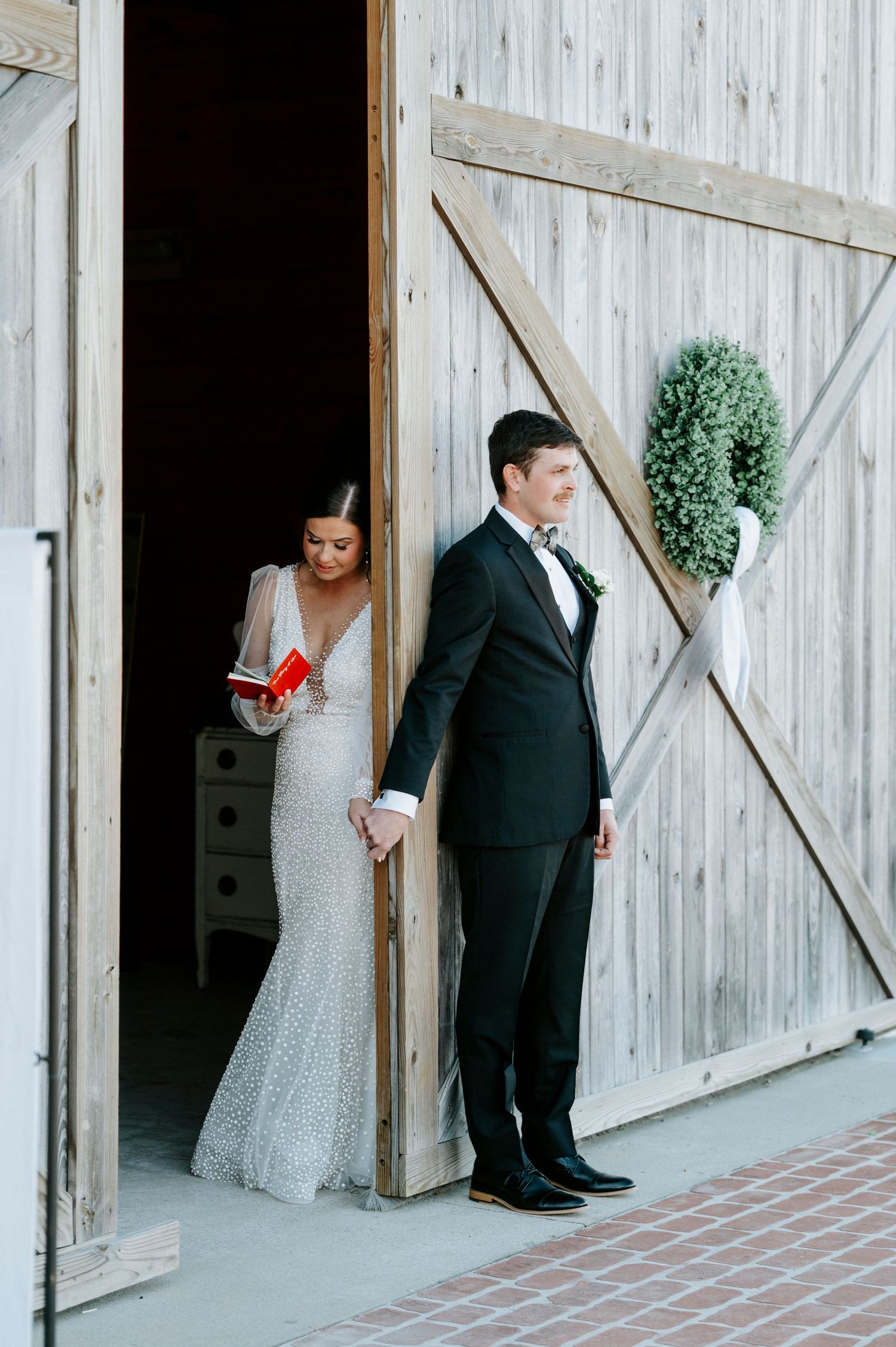 A rustic barn wedding scene with elegant formal attire and a decorative wreath on the white wooden door.