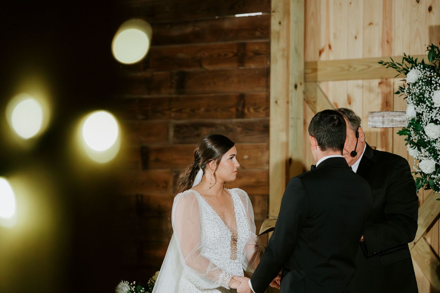 Inside a wooden barn venue, couple exchanges vows during intimate wedding ceremony with bokeh lights in foreground.