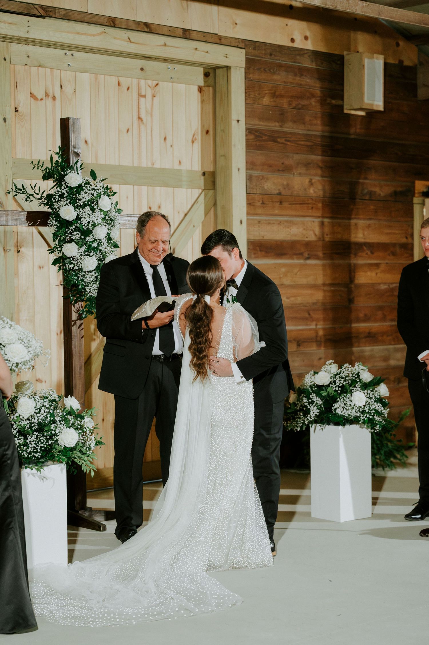 A wedding ceremony takes place in a rustic wooden venue decorated with elegant white floral arrangements.