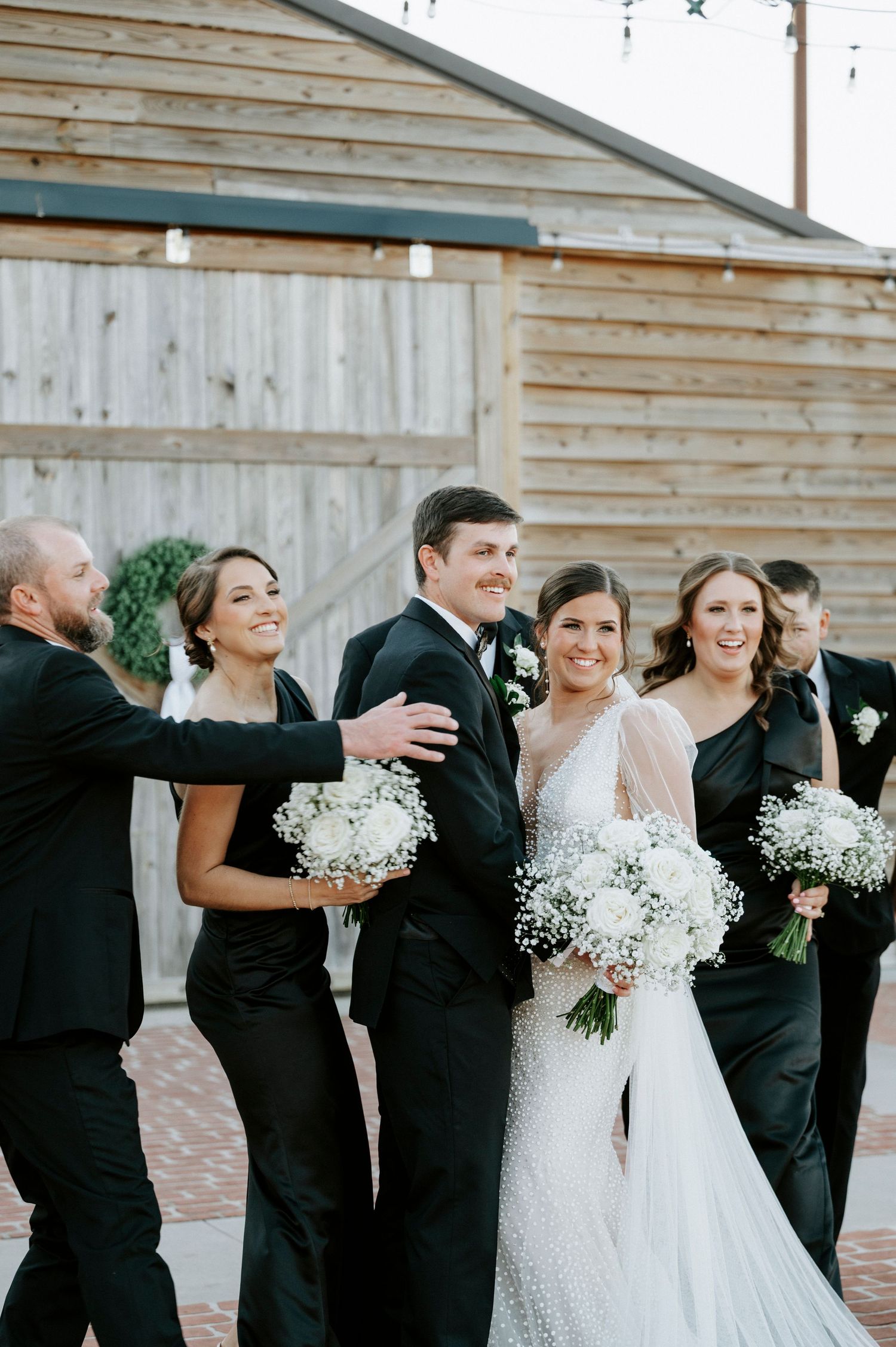 A wedding party in formal black attire and a white wedding gown celebrate together outdoors by a wooden building.