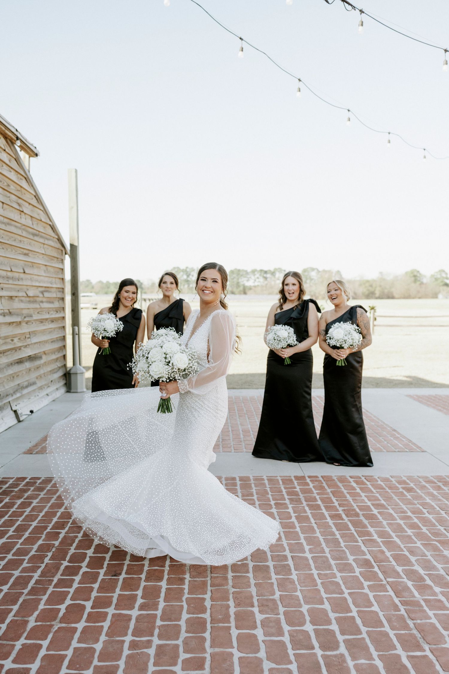 Wedding party in black dresses twirls and spins a flowing white wedding gown on a brick patio.