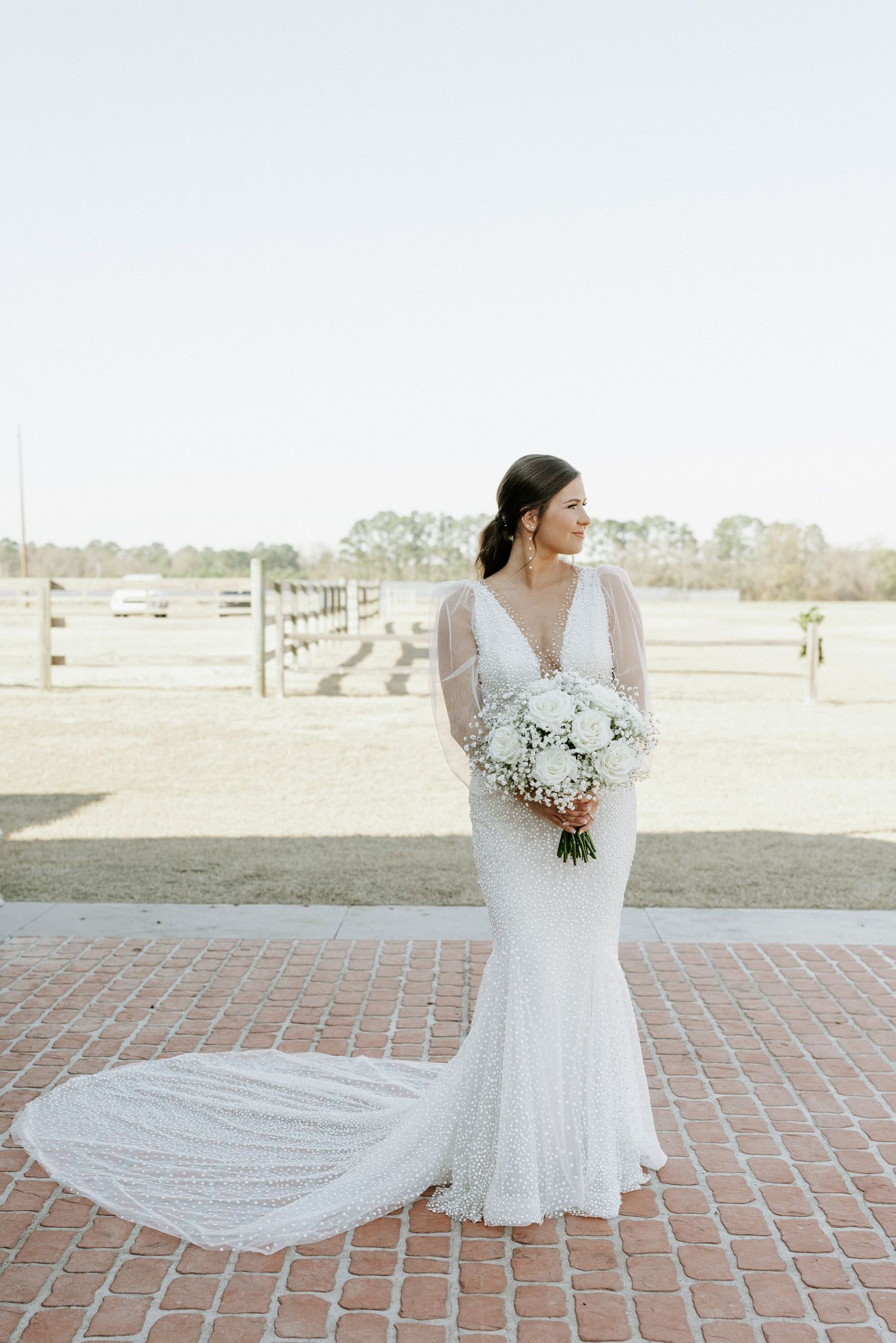 A bride in a fitted white gown with long train holds a white bouquet on a brick pathway.