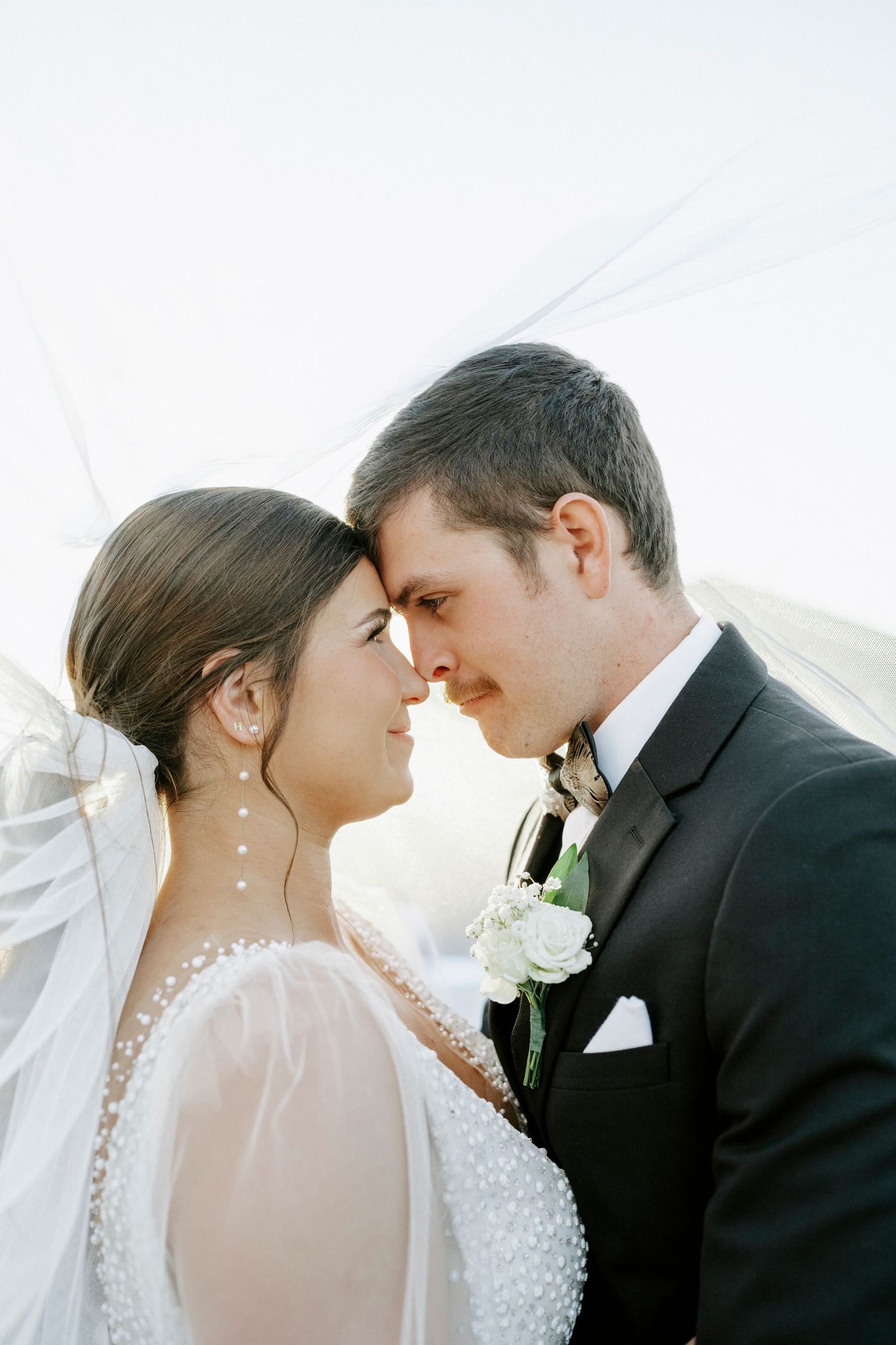 A romantic wedding portrait showing a couple in formal attire sharing an intimate moment against a bright background.