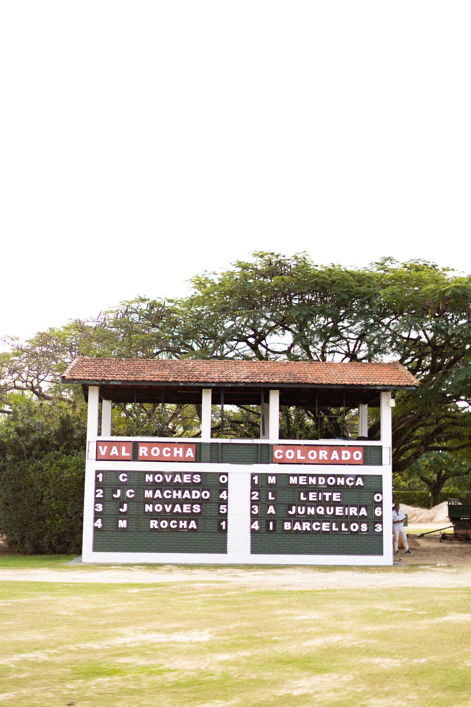 A vintage-style scoreboard display showing team names Val Rocha and Colorado under large shade trees.