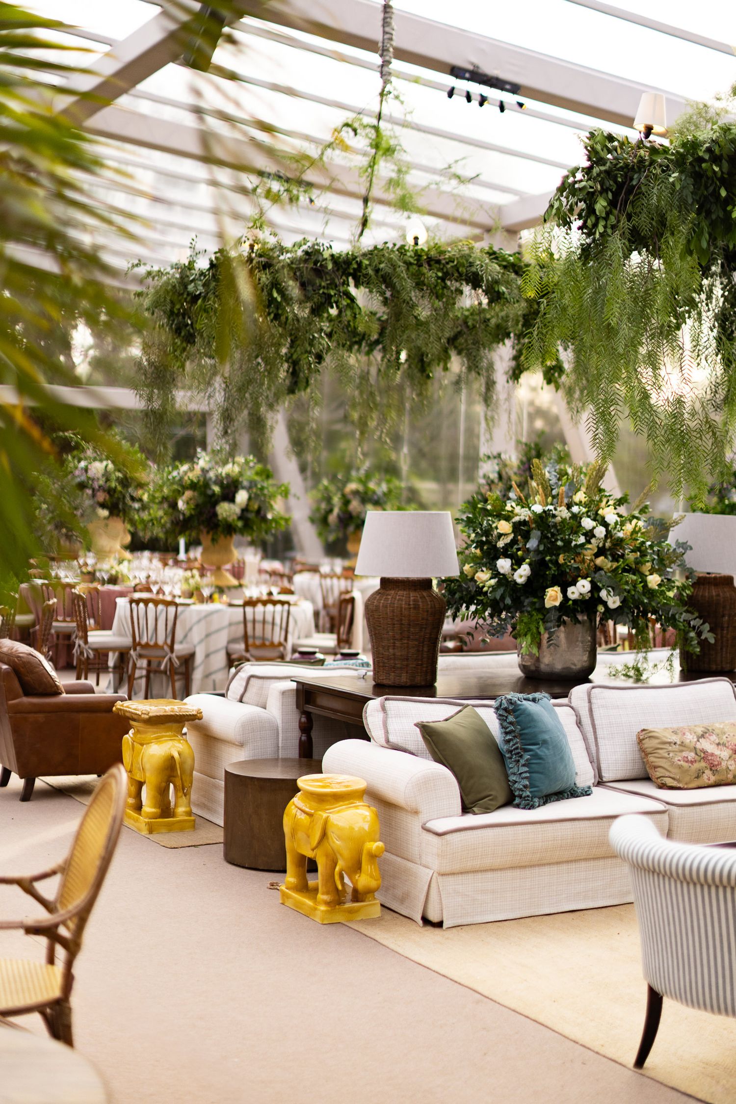 Elegant wedding reception lounge area with white sofas, hanging greenery, and yellow glass lanterns in a greenhouse setting.