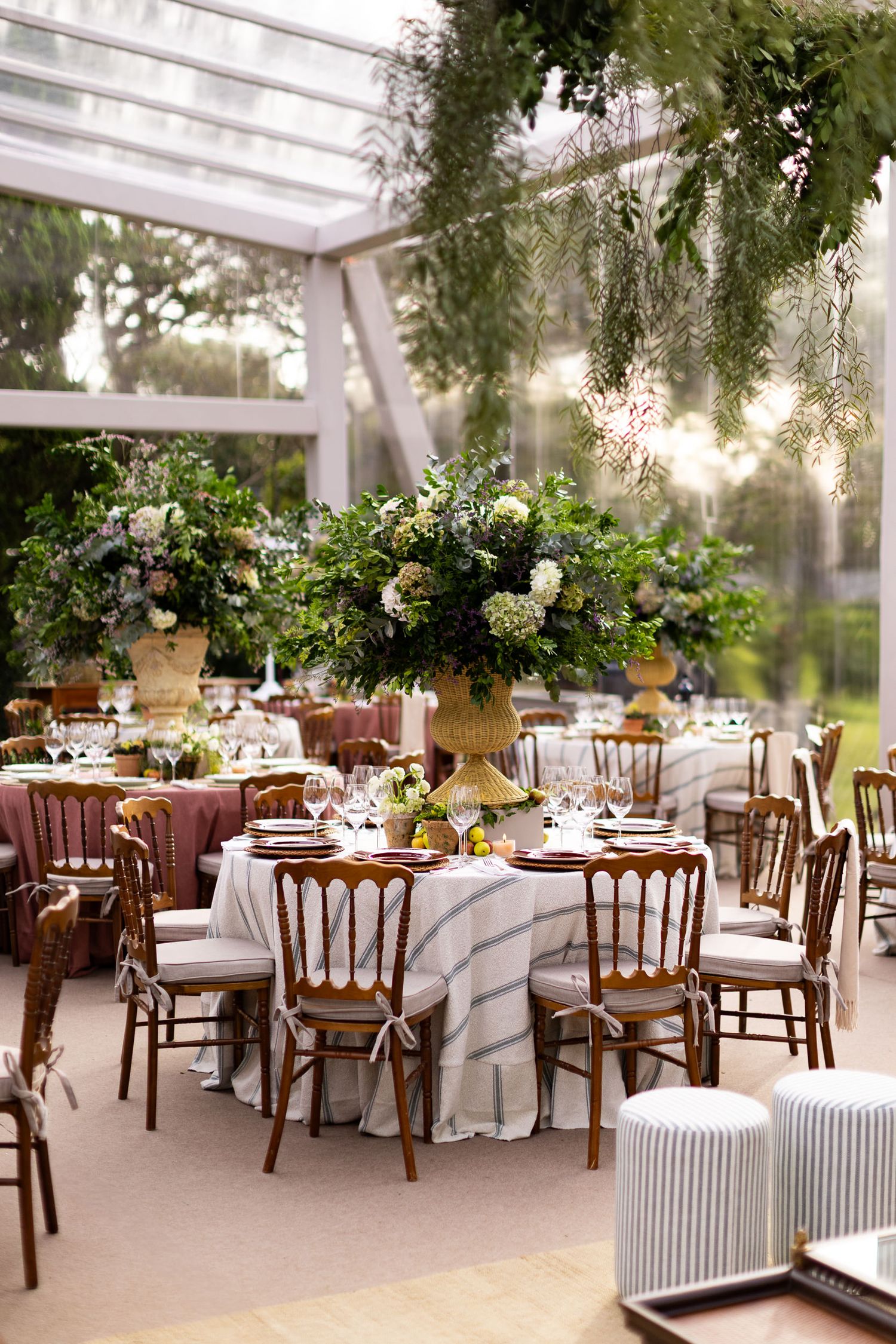 Romantic greenhouse wedding reception with round tables, wooden chairs and lush floral centerpieces under hanging greenery.