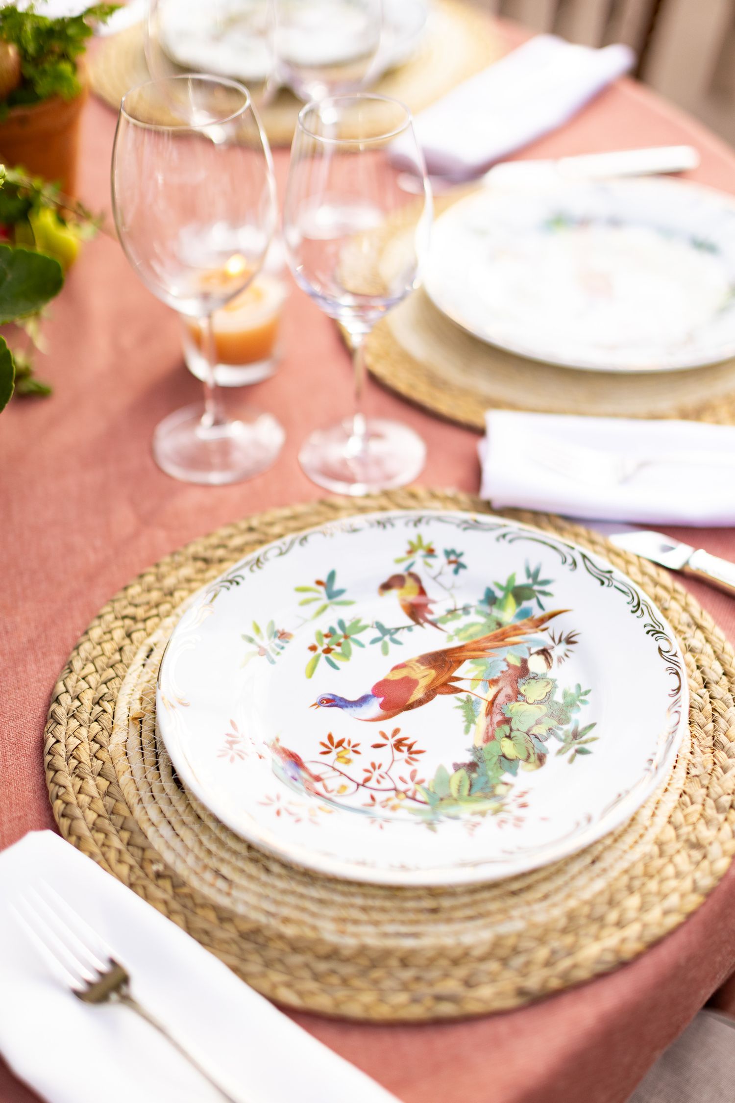 Elegant table setting with decorative china plate featuring bird motif on woven placemat with wine glasses and coral tablecloth.
