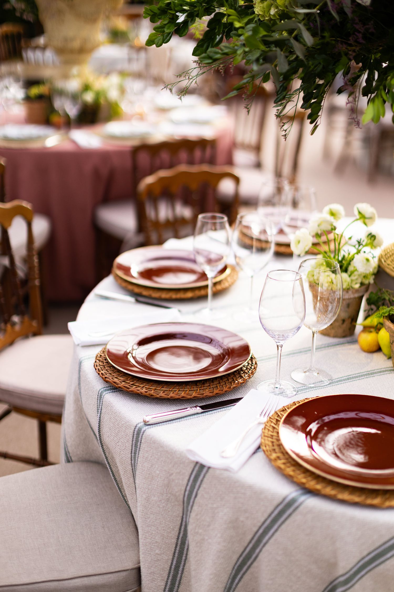 Elegant table settings with brown plates on woven chargers and white tablecloths decorated with fresh greenery.
