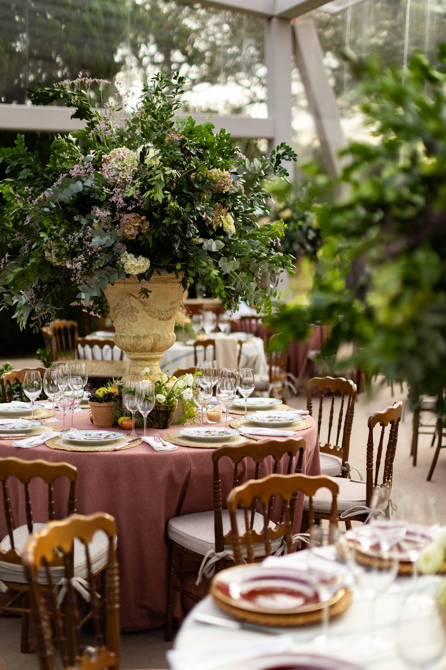 Elegant wedding reception tables set with burgundy linens, wooden chairs, and lush greenery centerpieces in a garden setting.