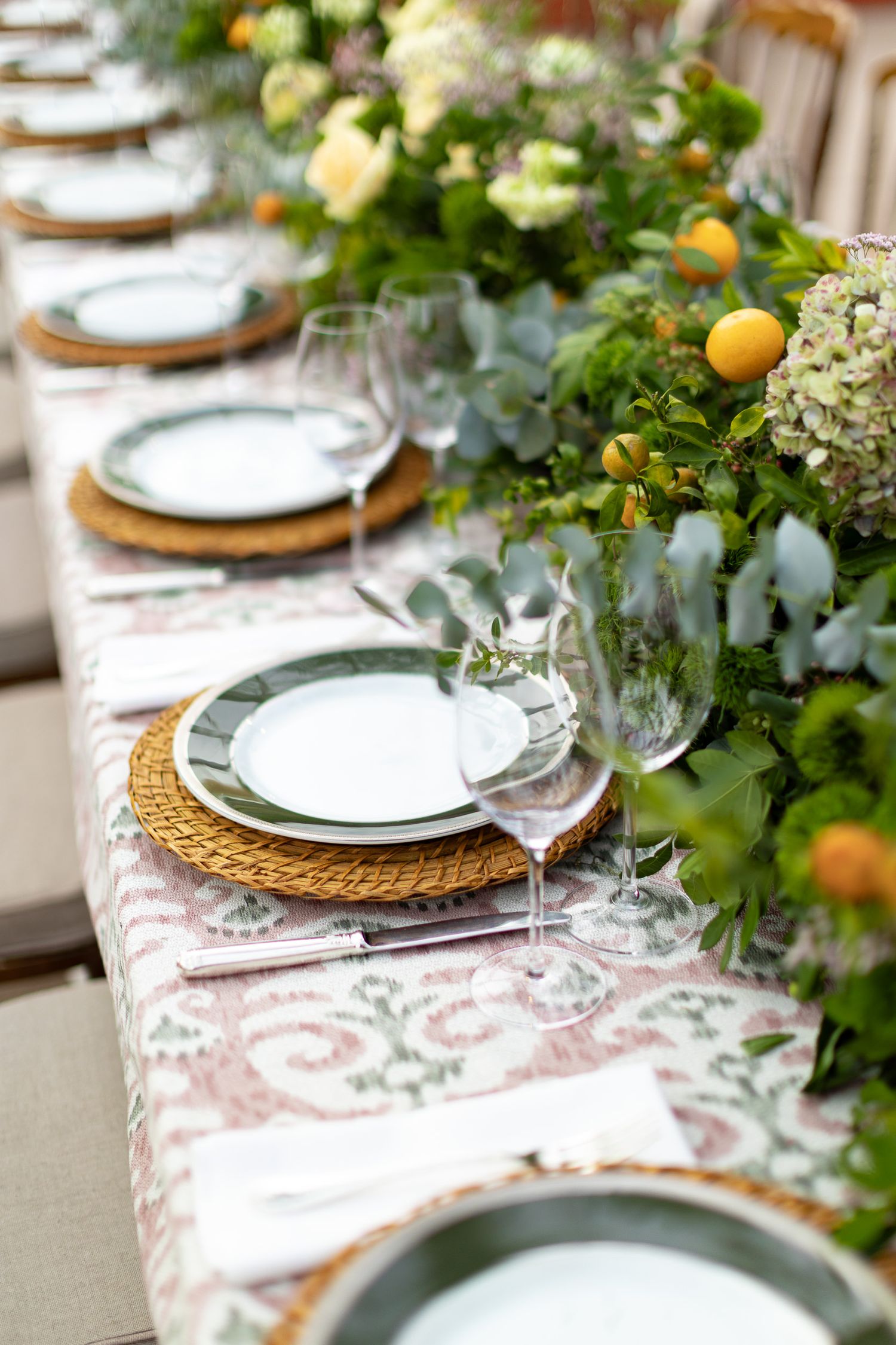 Formal table setting with white plates, woven chargers, and lush greenery centerpiece featuring citrus accents.