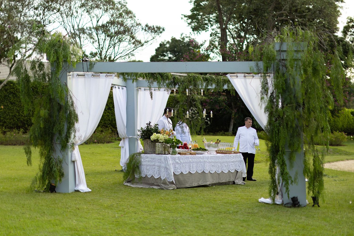 Elegant outdoor wedding reception setup with white draped canopy and decorated buffet table on a green lawn.