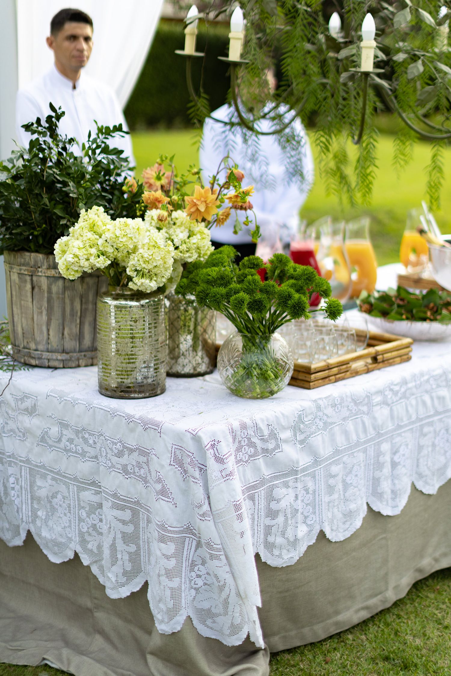 Elegant outdoor table setting with white lace tablecloth, glass vases of hydrangeas, and rustic decor in a garden setting.