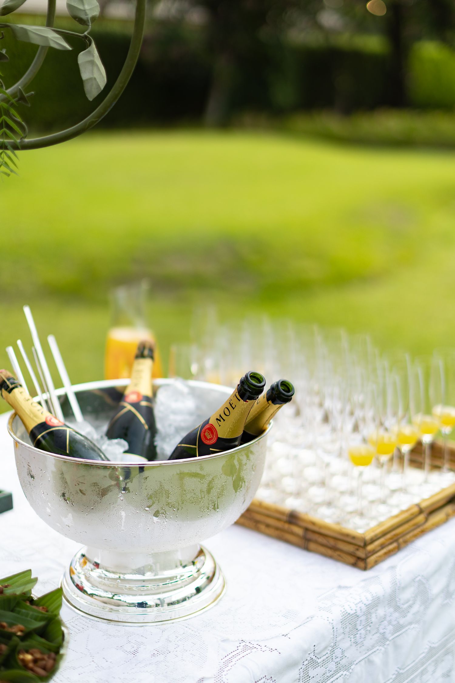 Champagne bottles chill in a silver ice bucket next to elegant glassware on a white tablecloth at an outdoor event.