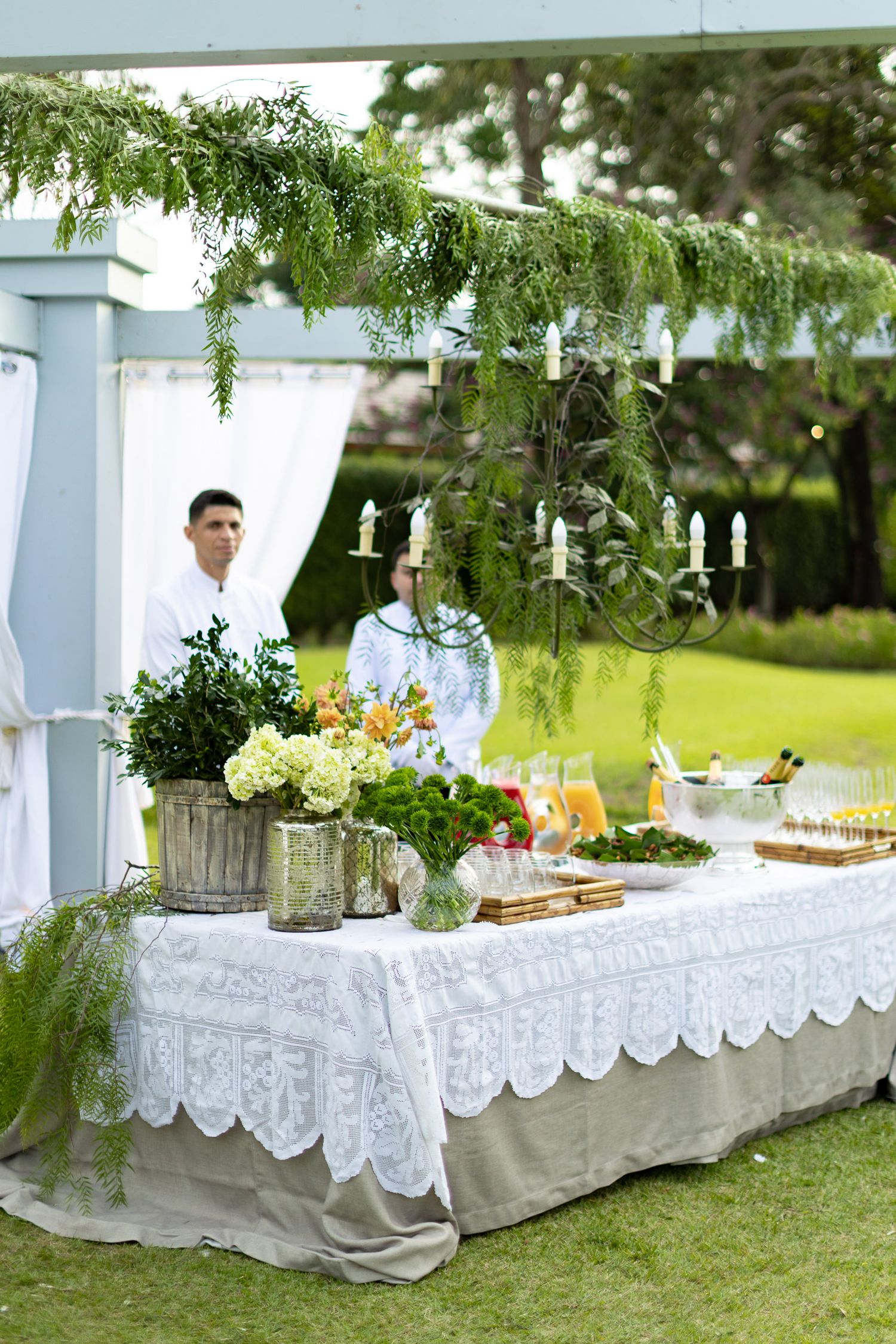 Elegant white lace tablecloths decorate outdoor buffet tables with drinks and refreshments at a garden party setting.