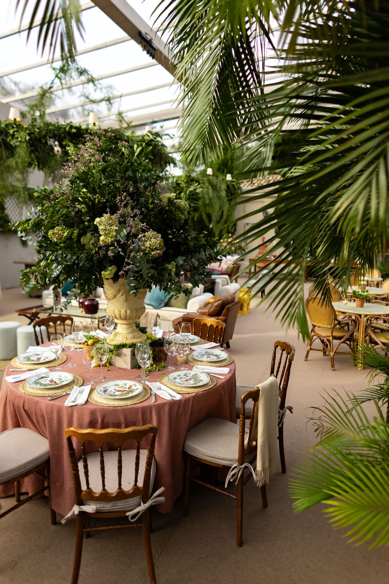 Elegant round dining tables with pink tablecloths set for a special occasion in a greenhouse-style venue with tropical plants.