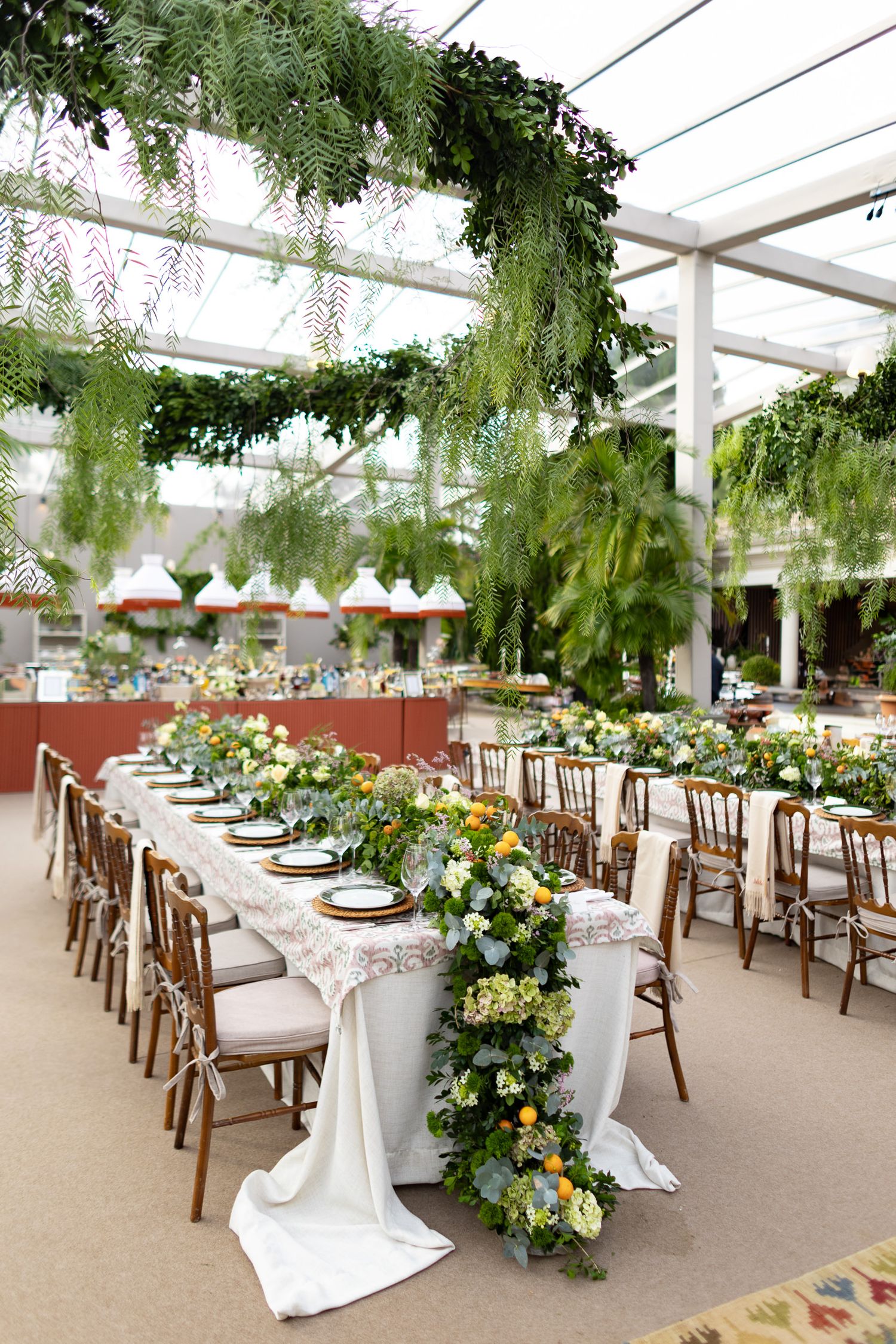 Wedding reception tent with long banquet tables decorated with lush greenery and floral arrangements under draped ceiling.