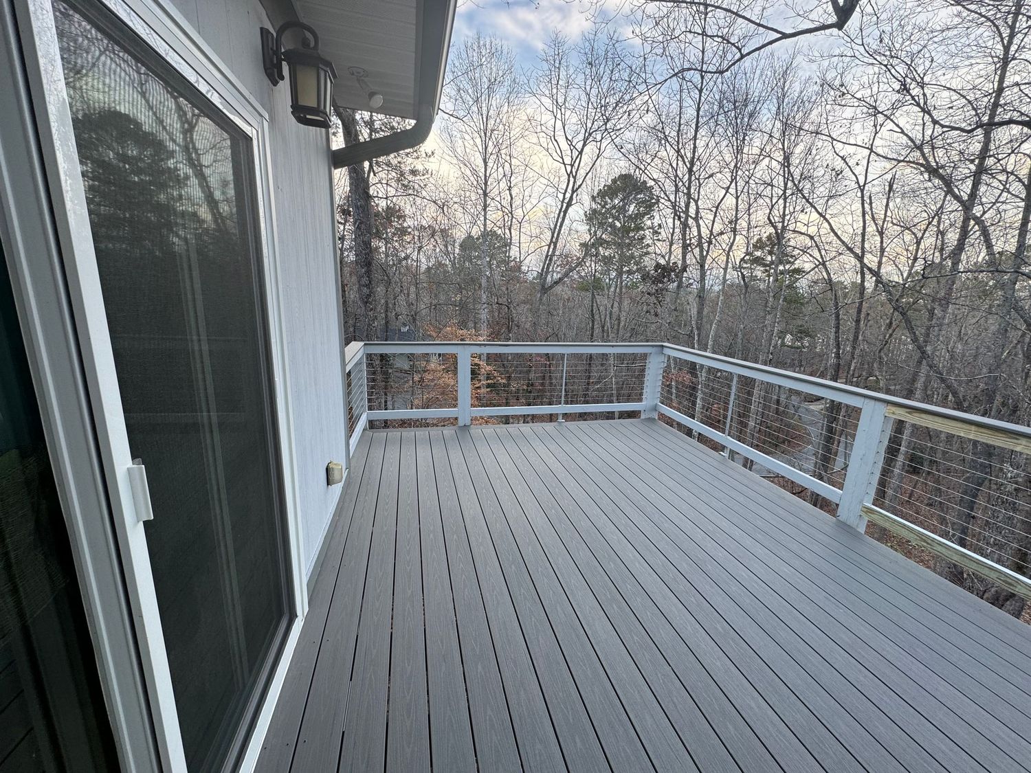 Modern gray composite deck with metal cable railings overlooks a winter forest landscape at dusk.