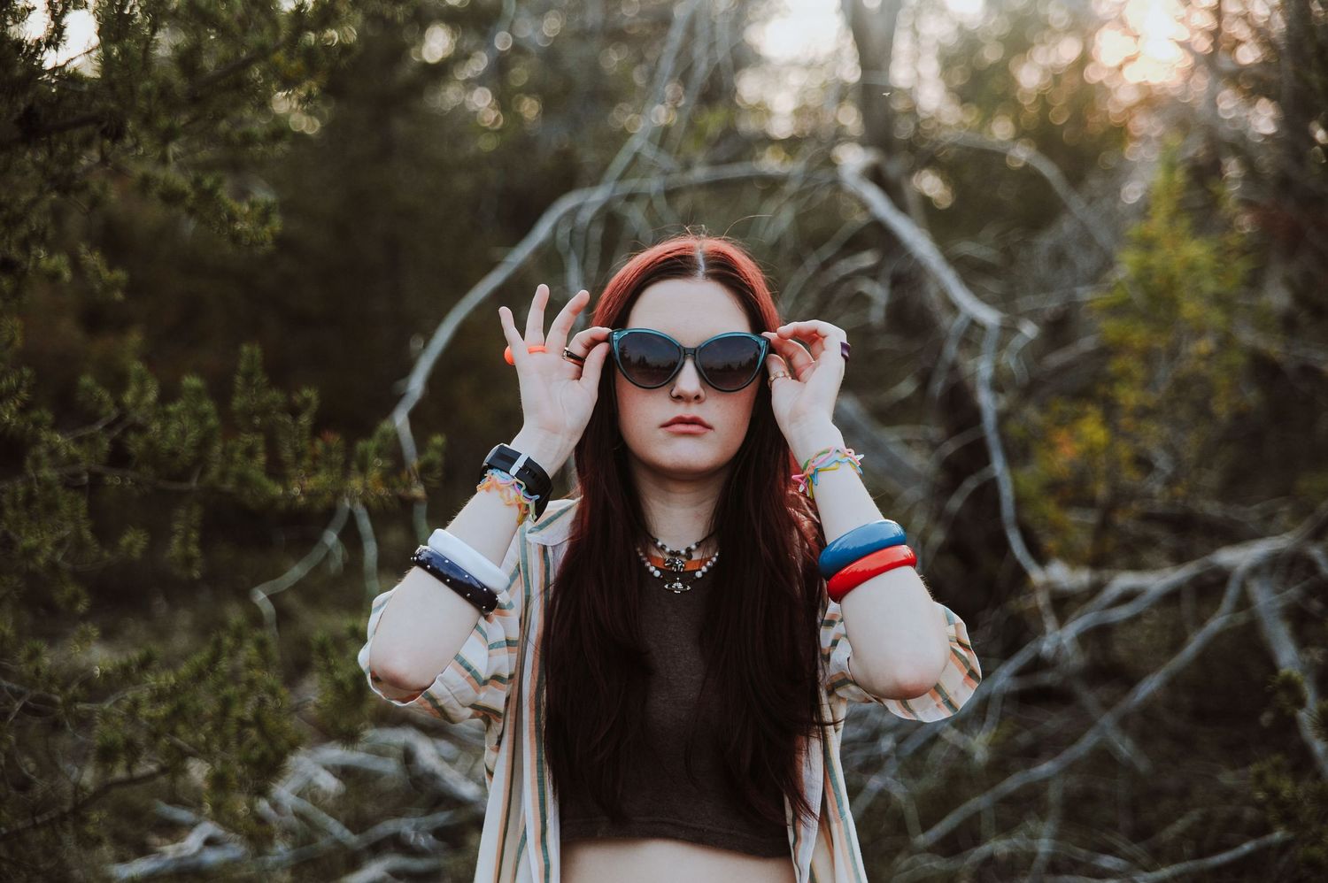 A person in sunglasses and a striped sweater poses in a forest setting during golden hour.
