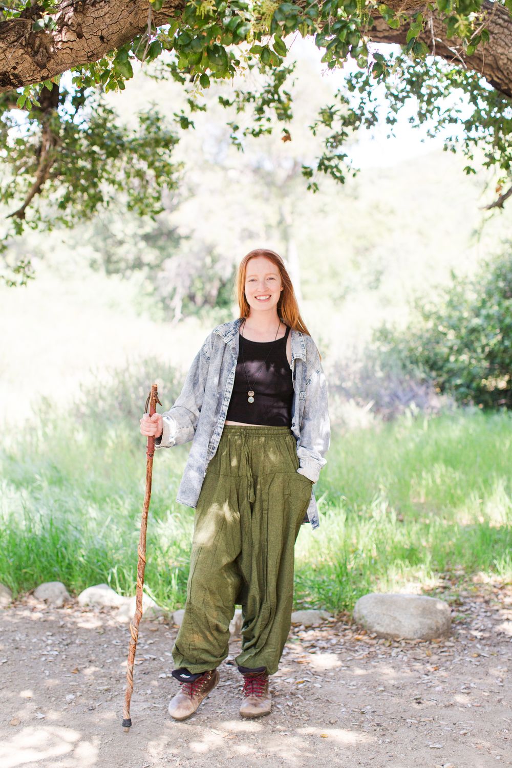 Person wearing olive pants and denim jacket holds walking stick in outdoor garden setting.