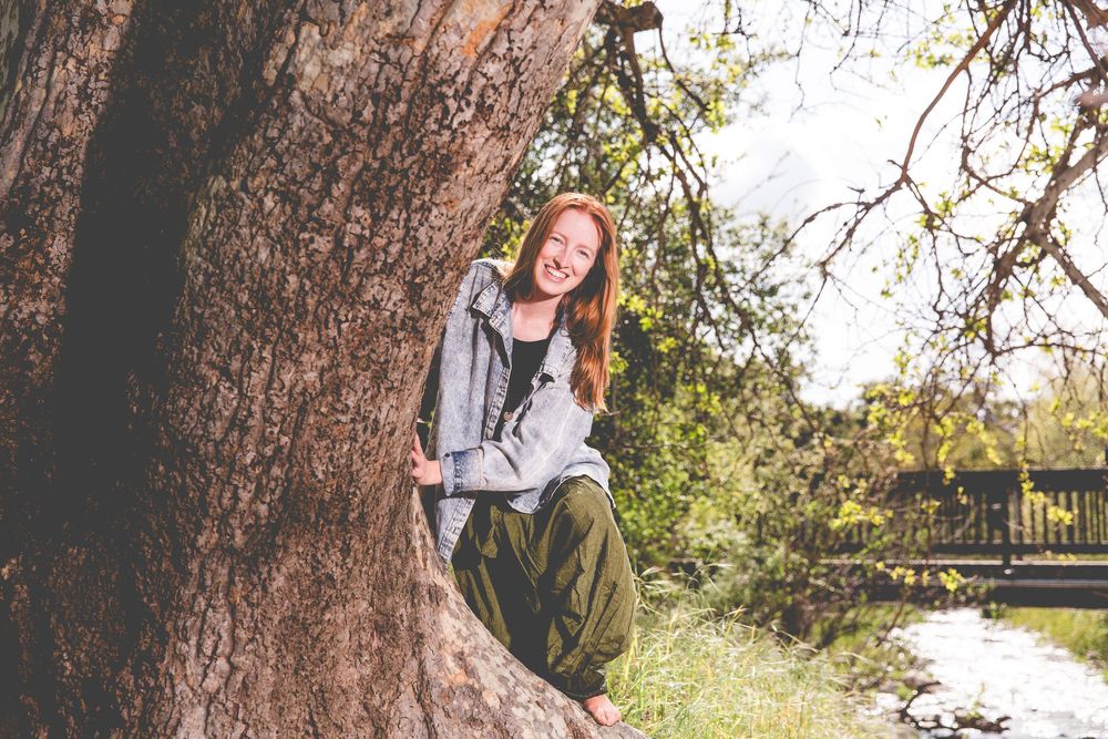Casual outdoor portrait against a rustic tree trunk in a sunny garden setting.