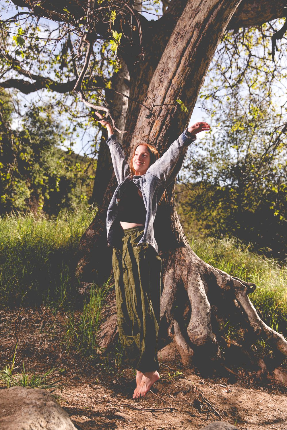 Joyful pose with arms raised against a large tree trunk in an outdoor natural setting.