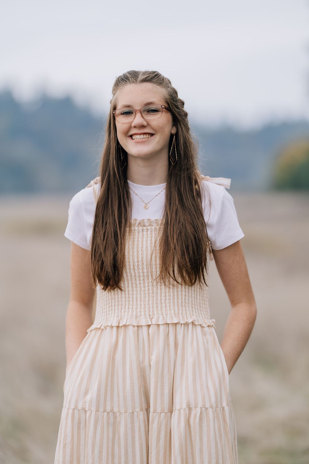 A smiling teenage girl with long brown hair and glasses poses in a striped sundress outdoors.