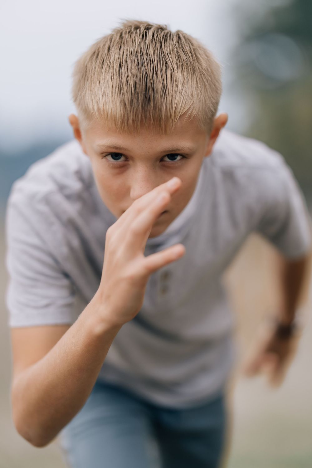 A blonde boy in a gray t-shirt leans forward gesturing with his hand toward the camera.