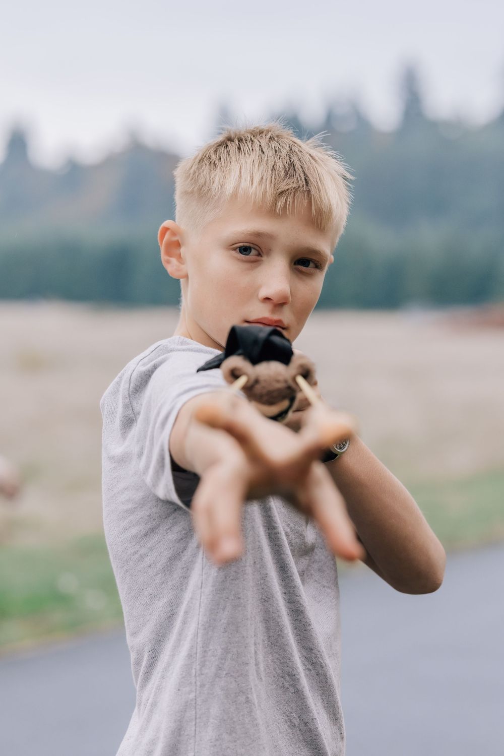 A young blonde boy in a gray t-shirt points a toy gun toward the camera outdoors.