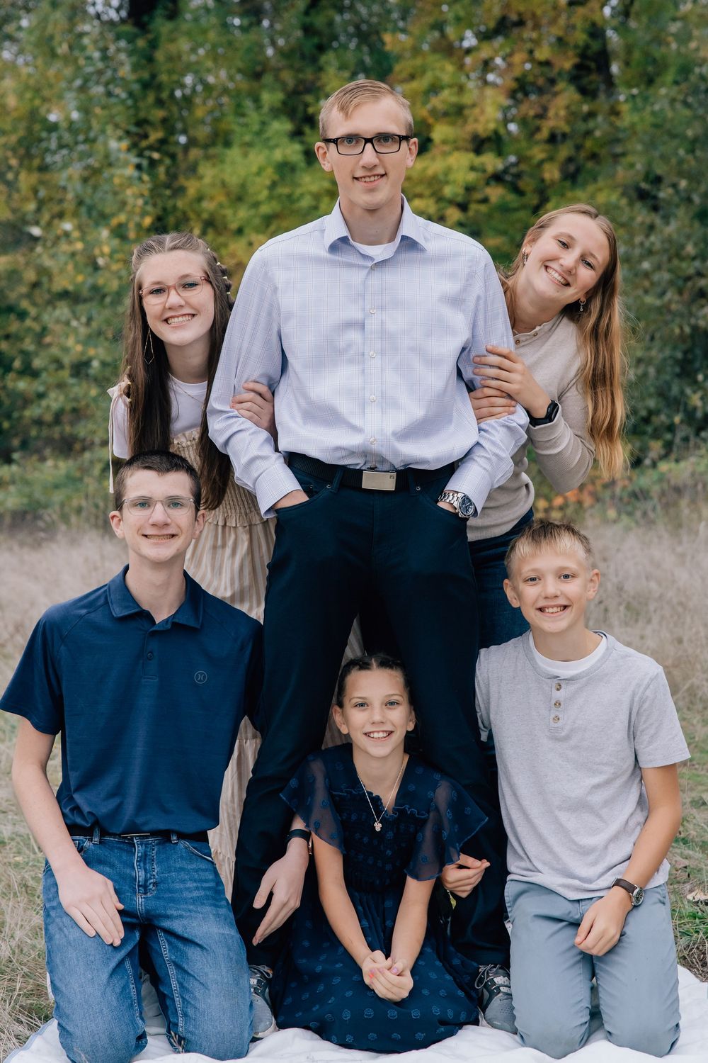 Six siblings pose together in a playful pyramid formation in an outdoor field setting.