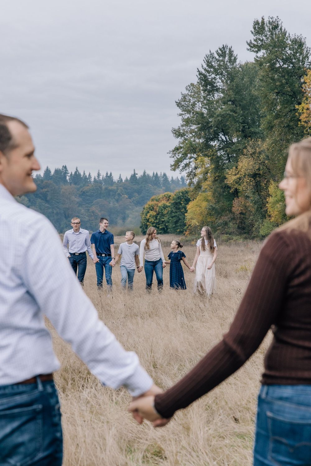 A couple holds hands in a field while their children walk toward them in the background.