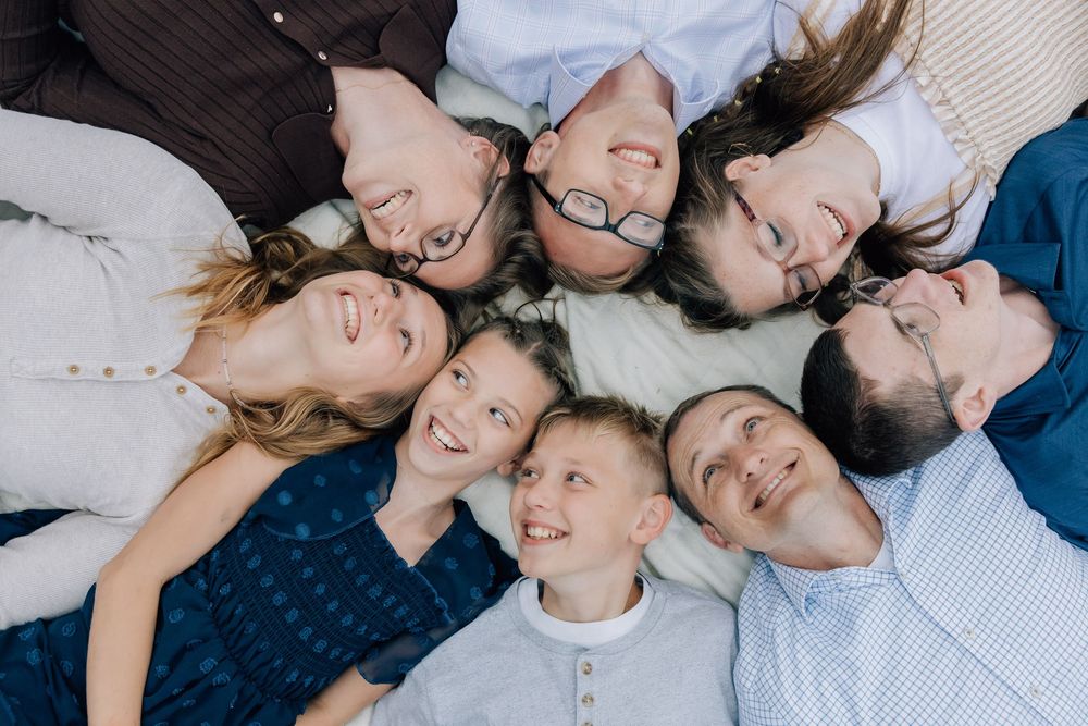 A happy family of seven lies in a circle on the ground smiling up at the camera.