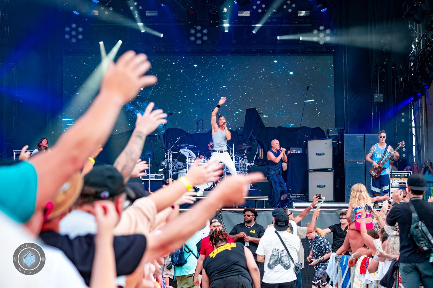 Energetic outdoor concert crowd raising hands toward performer on stage with band and speakers behind.