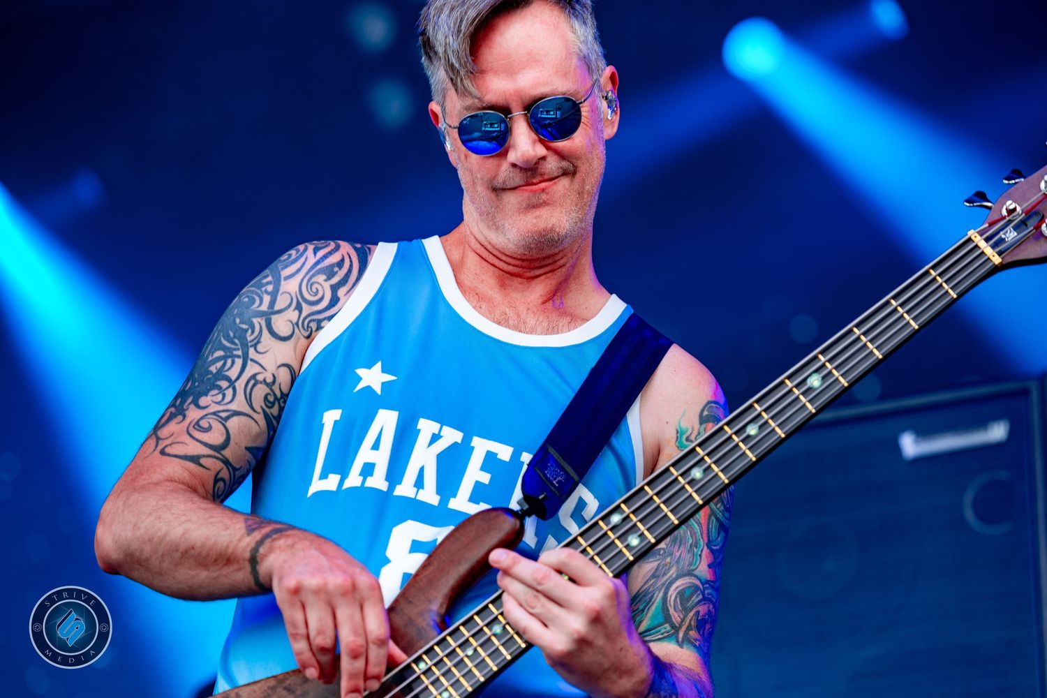 Tattooed musician 311 playing bass guitar on stage, wearing blue Lakers jersey and sunglasses under blue stage lighting.