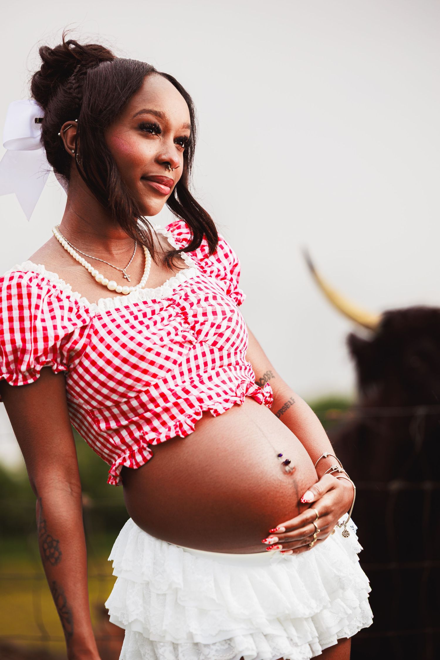 Smiling pregnant Black woman in red gingham crop top and white skirt posing outdoors near a bull.