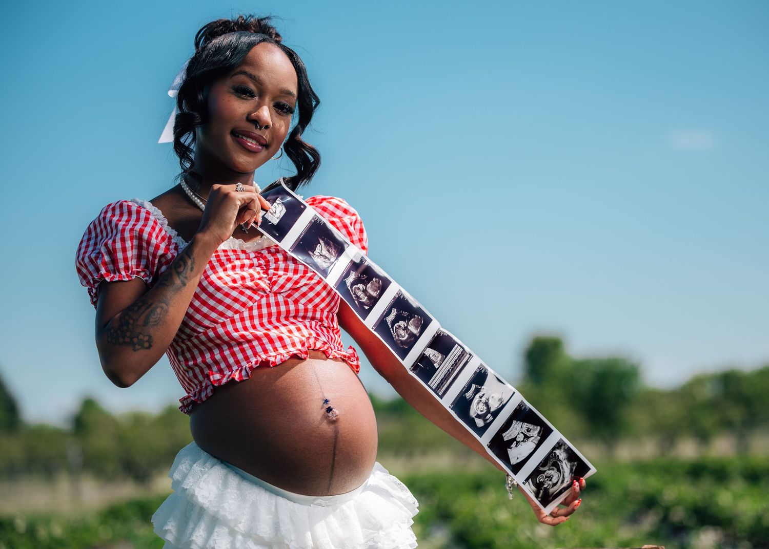 Happy pregnant woman in red gingham top holding ultrasound strip photos outdoors in a field.