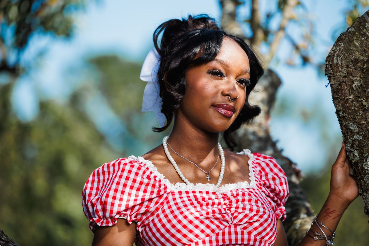 Beautiful Black woman in red gingham dress leaning against a tree in a sunny outdoor setting.