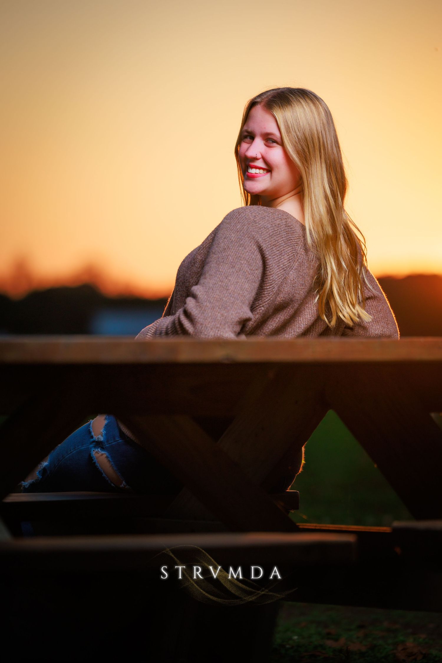 Young blonde woman sitting on a wooden bench smiling over her shoulder during a golden sunset outdoors.