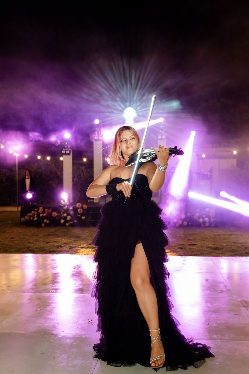A violinist performs in a dramatic black gown with purple stage lighting and fog effects at an outdoor event.